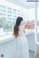 A woman in a white dress standing by a window.