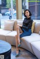 A woman sitting on a couch in a black shirt and blue skirt.