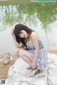 A woman in a gray dress sitting on a rock by the water.