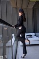 A woman in a black top and black pants leaning against a glass railing.